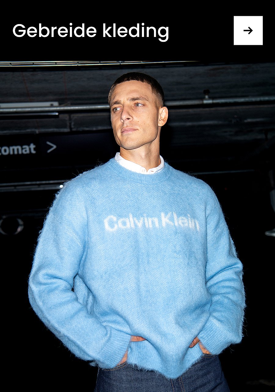 Een glimlachende man in een blauwe trui en een grijze broek, zittend op een rood tapijt. Tekst: "Broek".