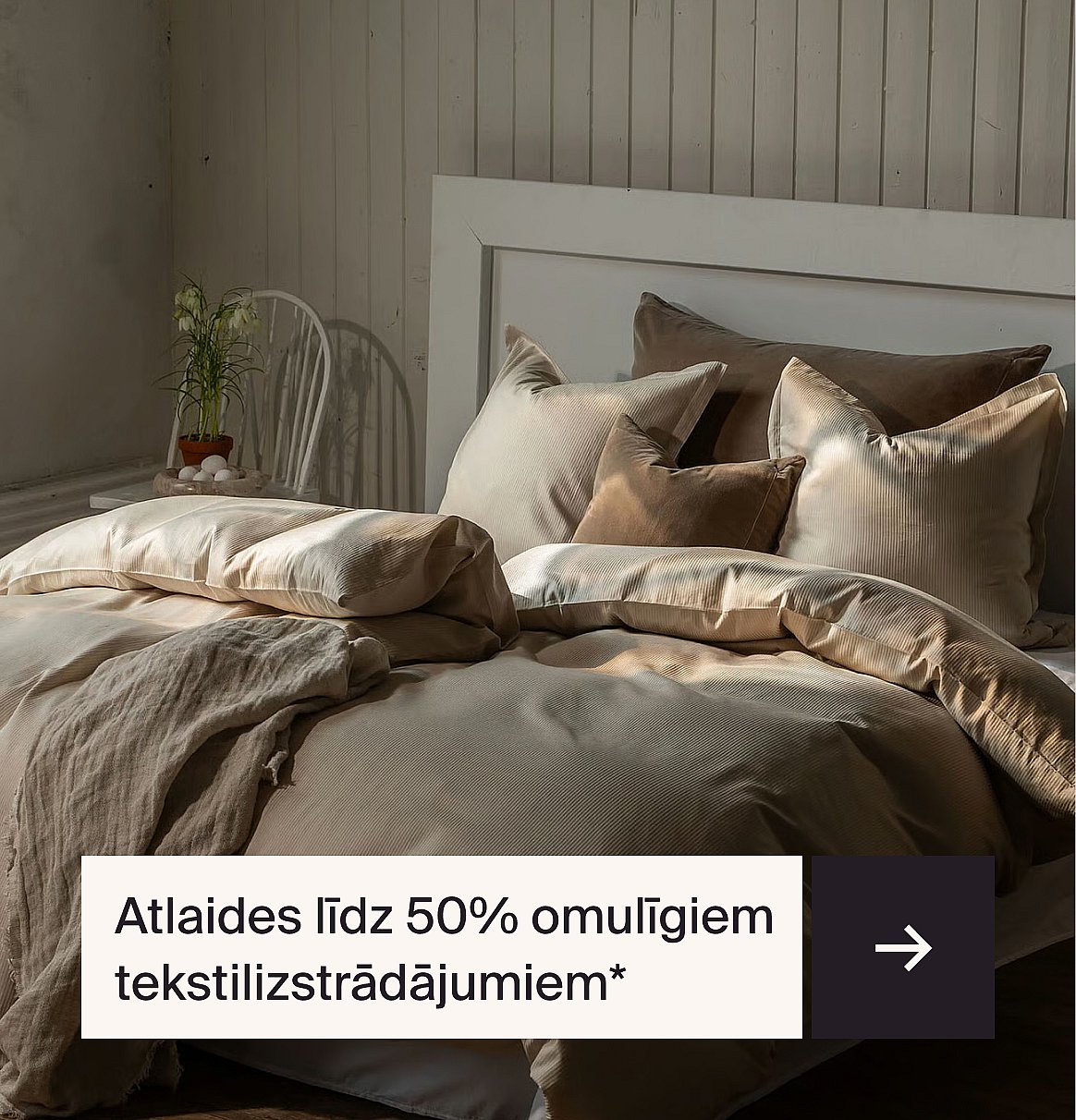 Mājīga gulta ar smilškrāsas un brūnas velveta gultas veļu, baltu galvgalvi un augu uz krēsla. Teksts: Līdz pat 40% atlaide mājīgiem tekstilizstrādājumiem.