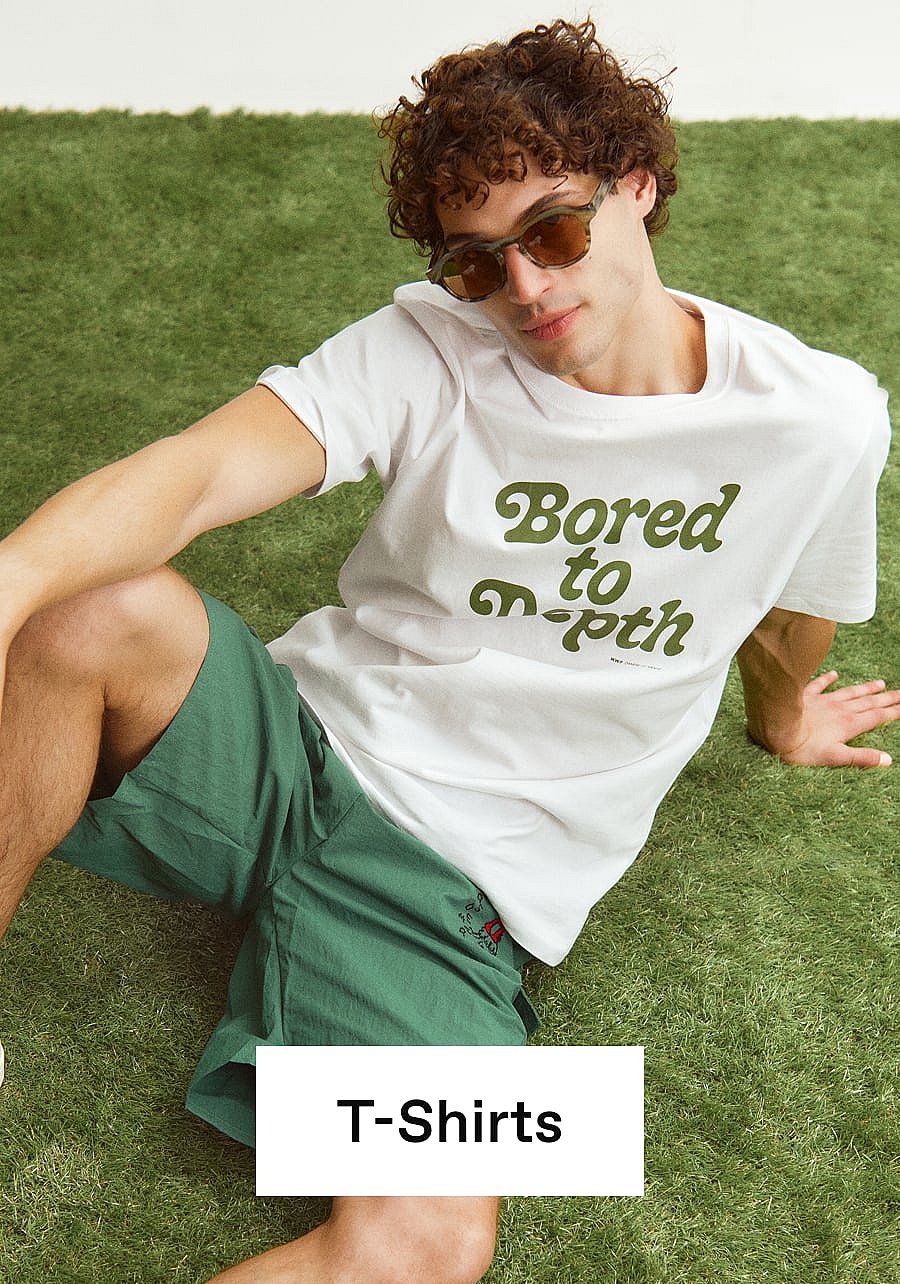 Mann mit lockigem Haar und Sonnenbrille sitzt auf grünem Gras und trägt ein weißes T-Shirt mit dem Aufdruck „Bored to Death“ sowie grüne Shorts.