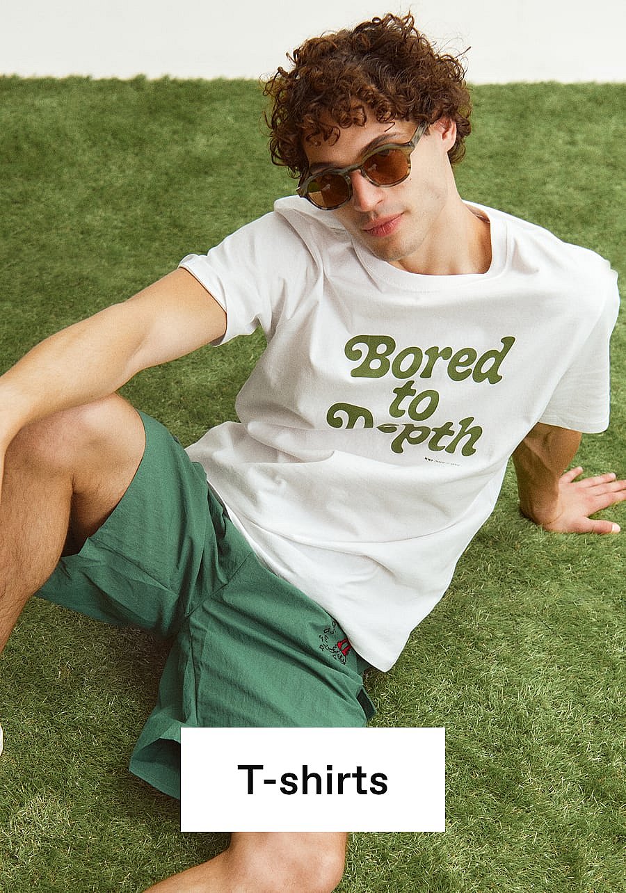 Man met krullend haar en zonnebril, zittend op groen gras, in een wit T-shirt met "Bored to Death" en groene korte broek.