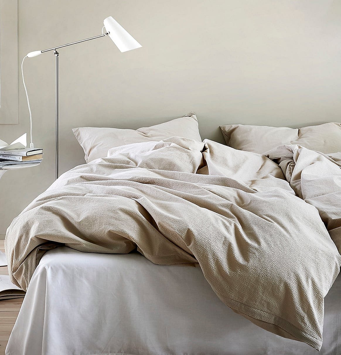 Ein Bett mit zerwühlter beiger Bettwäsche und Kissen, weissen Leintüchern und einer modernen Stehlampe in einem hellen Schlafzimmer.