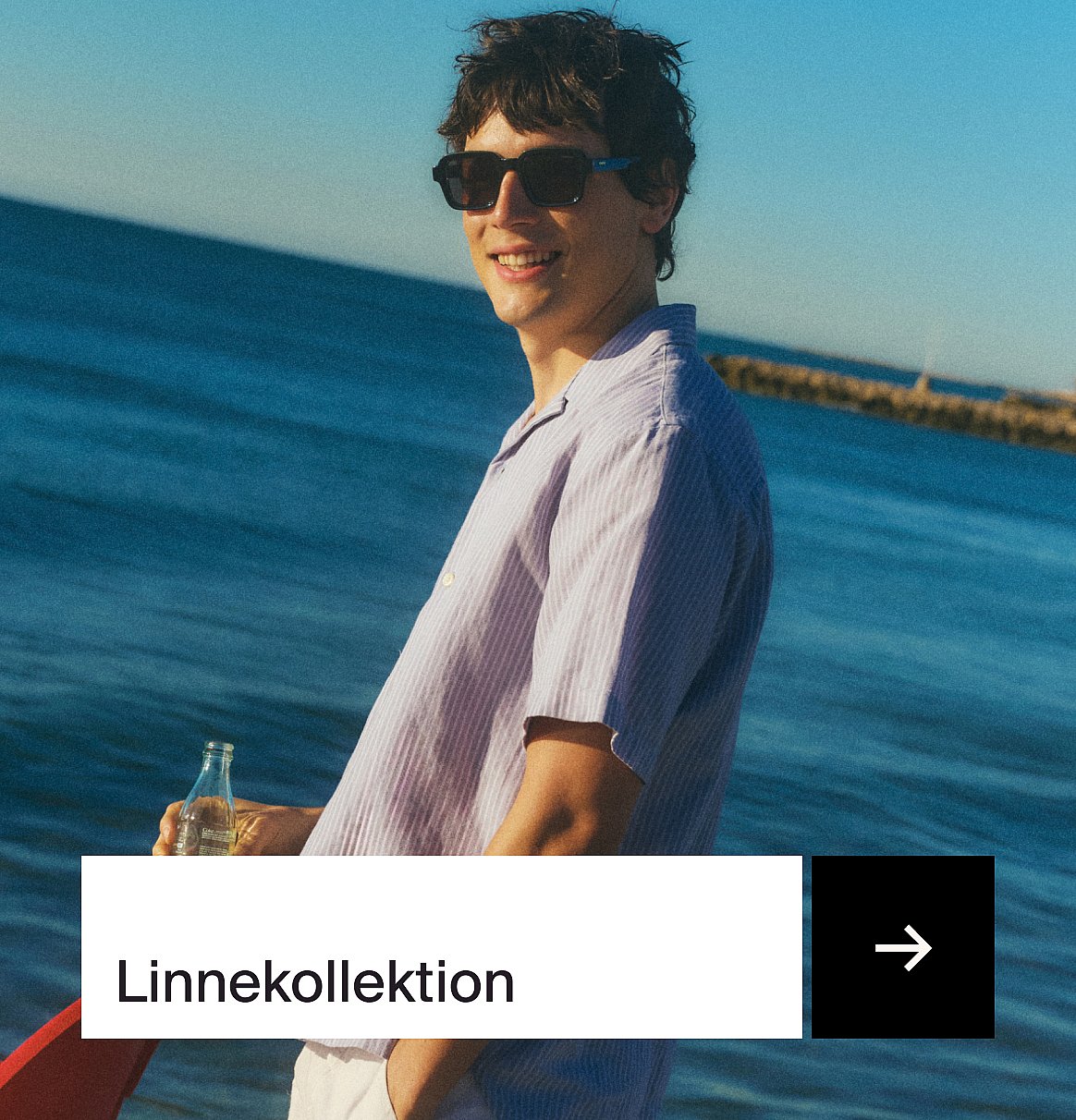 Leende man i lila-randig linneskjorta och solglasögon vid havet, hållande en flaska. Text: Linnekollektion.