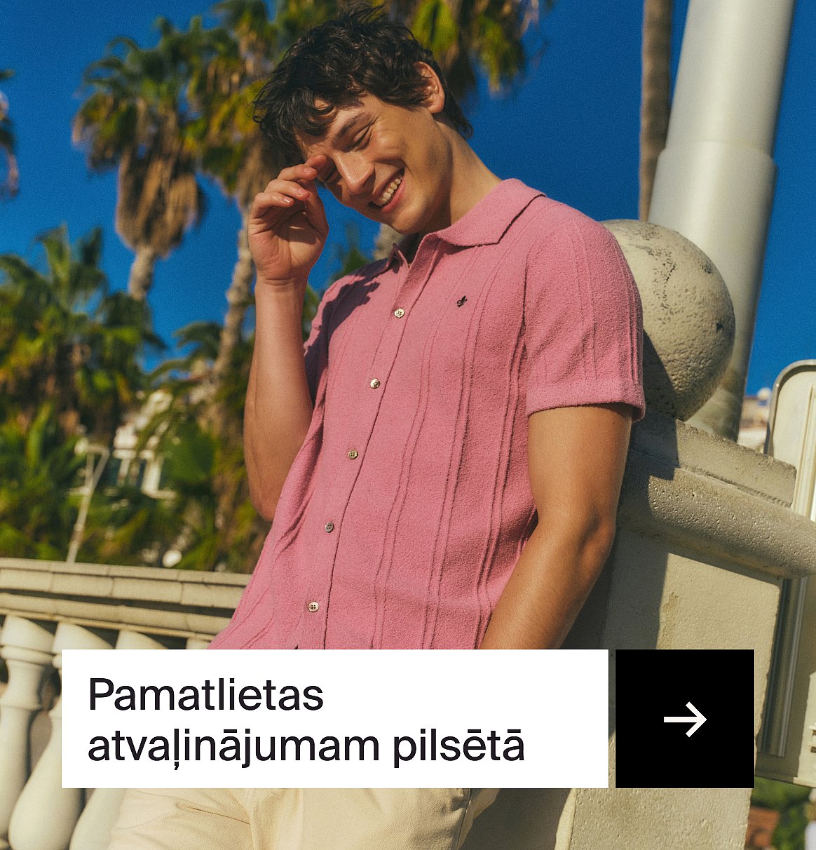 Smaidošs vīrietis rozā polo kreklā, ar roku pie sejas, atspiedies uz akmens balustrādes. Palmas un zilas debesis.