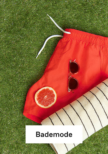Rote Badeshorts, Sonnenbrille, Grapefruitscheibe und gestreiftes Handtuch auf grünem Gras.
Bademode.