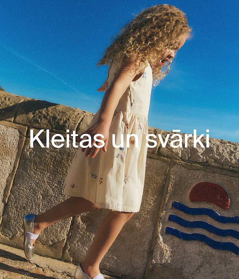 Cirtainu matu meitene krēmkrāsas ziedu kleitā, ejot garām akmens sienai zem koši zilām debesīm.
Teksts: Kleitas un svārki.