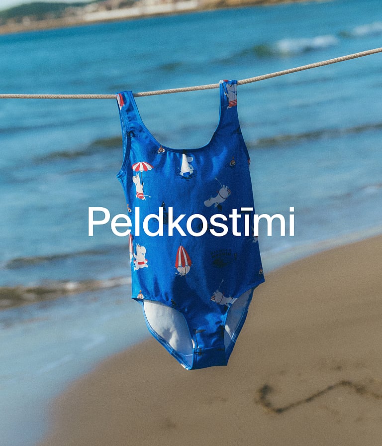 Zils Mūminu peldkostīms, piekārts uz veļas auklas virs pludmales ar okeāna viļņiem.
Teksts: Peldkostīmi.
