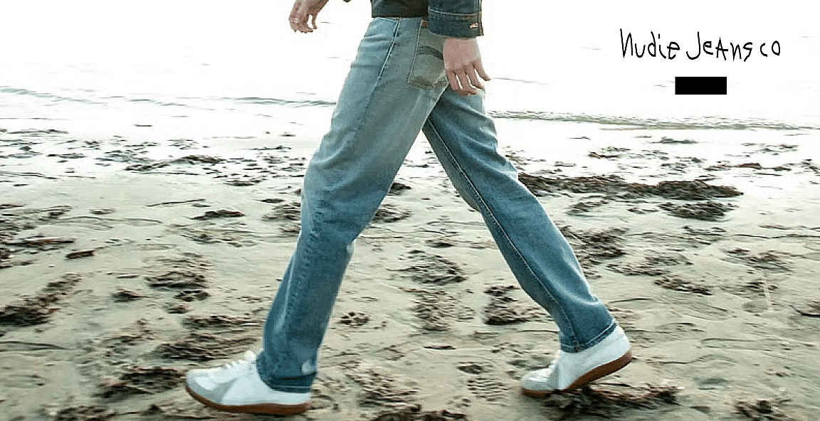 Tyrkise stribede badeshorts hænger på en tørresnor, med en solrig strand og hav i baggrunden. Tekst: Shop badeshorts.