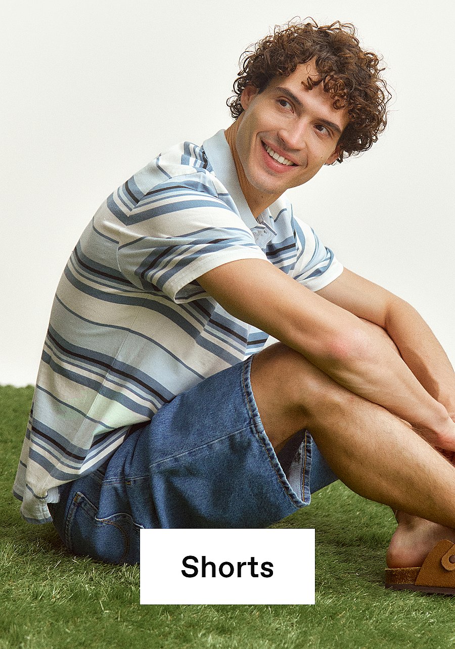 Lächelnder Mann mit lockigem Haar, in gestreiftem Poloshirt und Jeansshorts, auf grünem Gras sitzend.