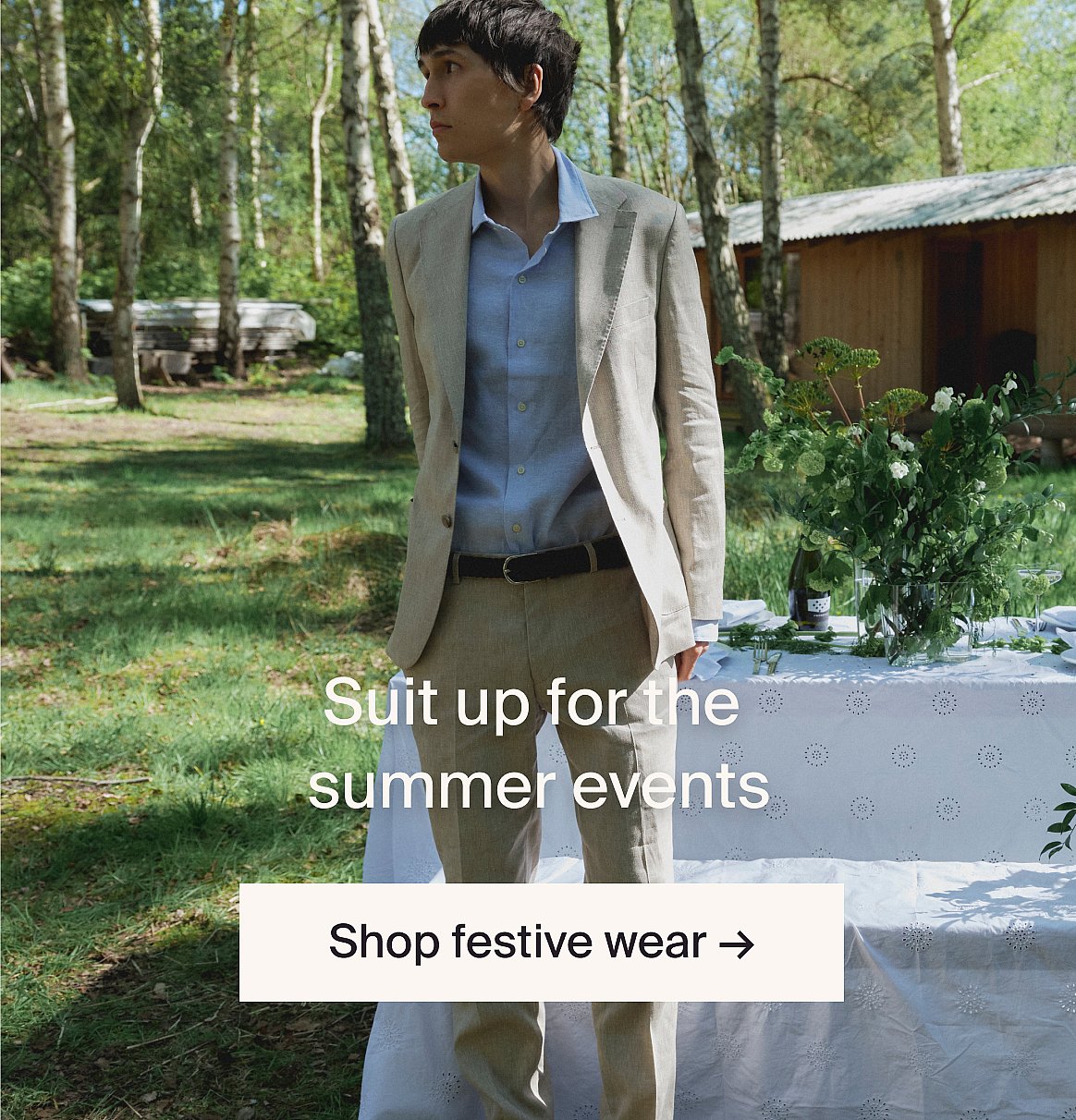 Man in een lichtbeige pak en blauw overhemd buiten, bij een tafel met groen, op een open plek in het bos.