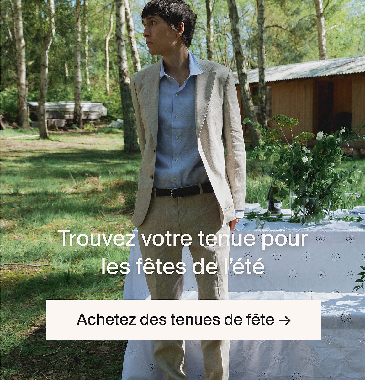 Homme en costume beige clair et chemise bleue en extérieur, près d'une table avec de la verdure, dans une clairière en forêt.