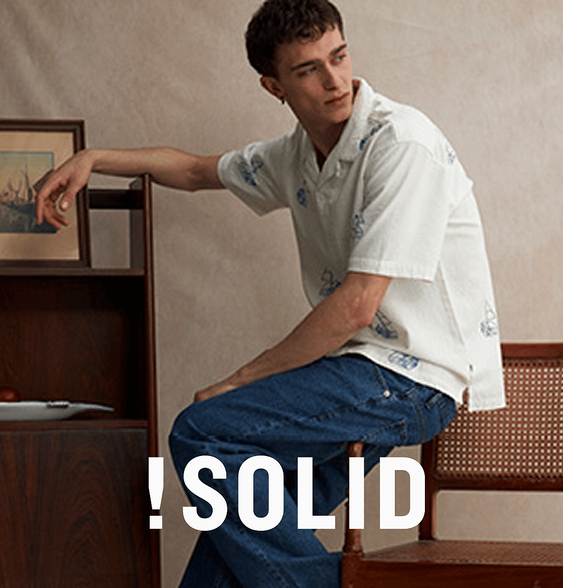 Person i vit kortärmad broderie anglaise-skjorta och blå jeans, händerna i fickorna, står mot en ljus trävägg.