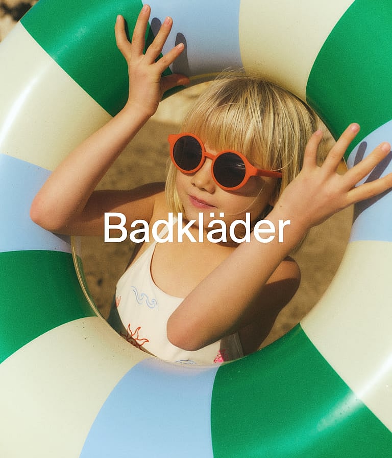 Blå Muminbaddräkt hängande på en tvättlina över en strand med havsvågor. Text: Badkläder.