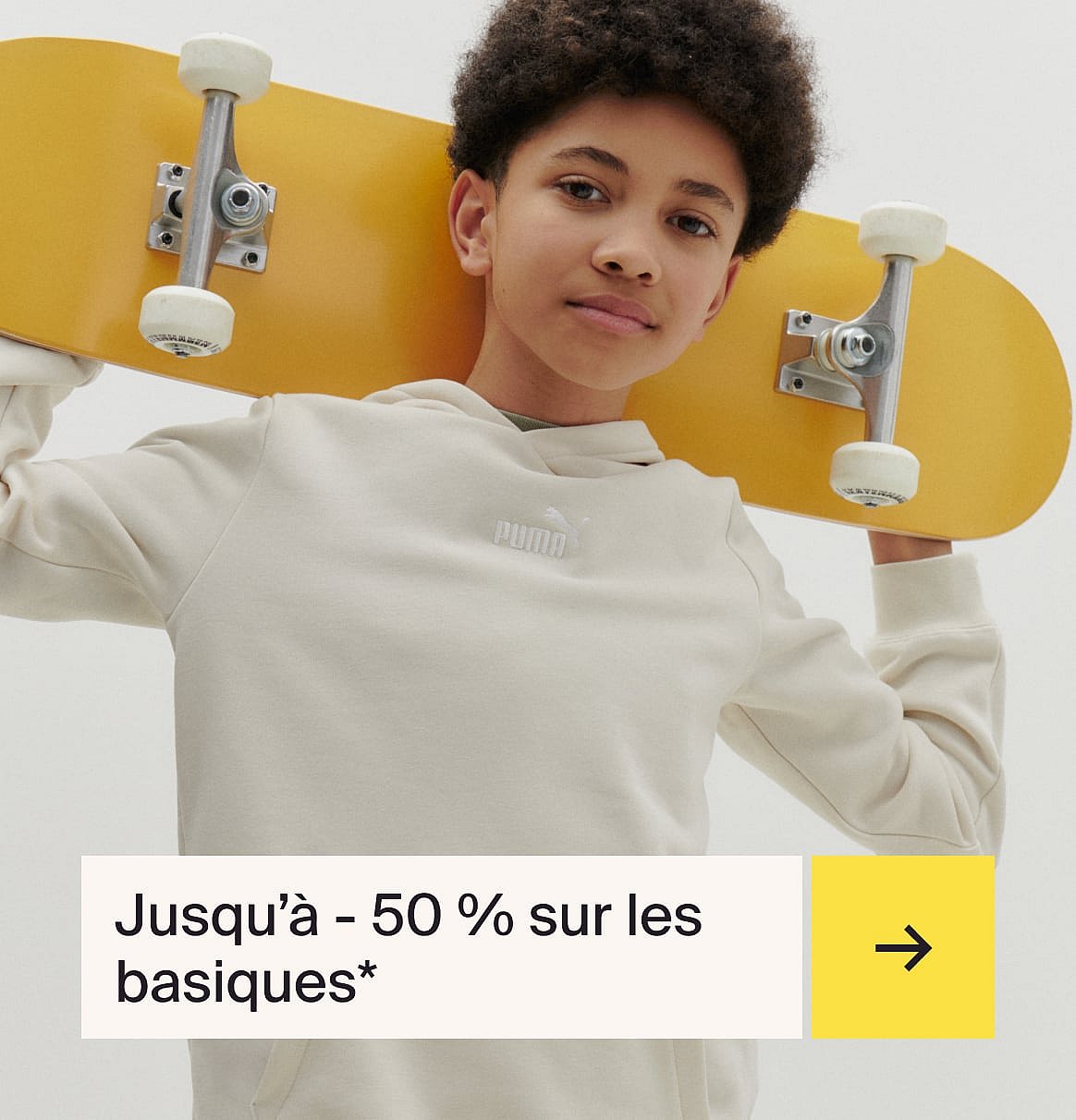 Young person with curly hair wearing a beige Puma hoodie, holding a yellow skateboard behind their neck, looking forward.