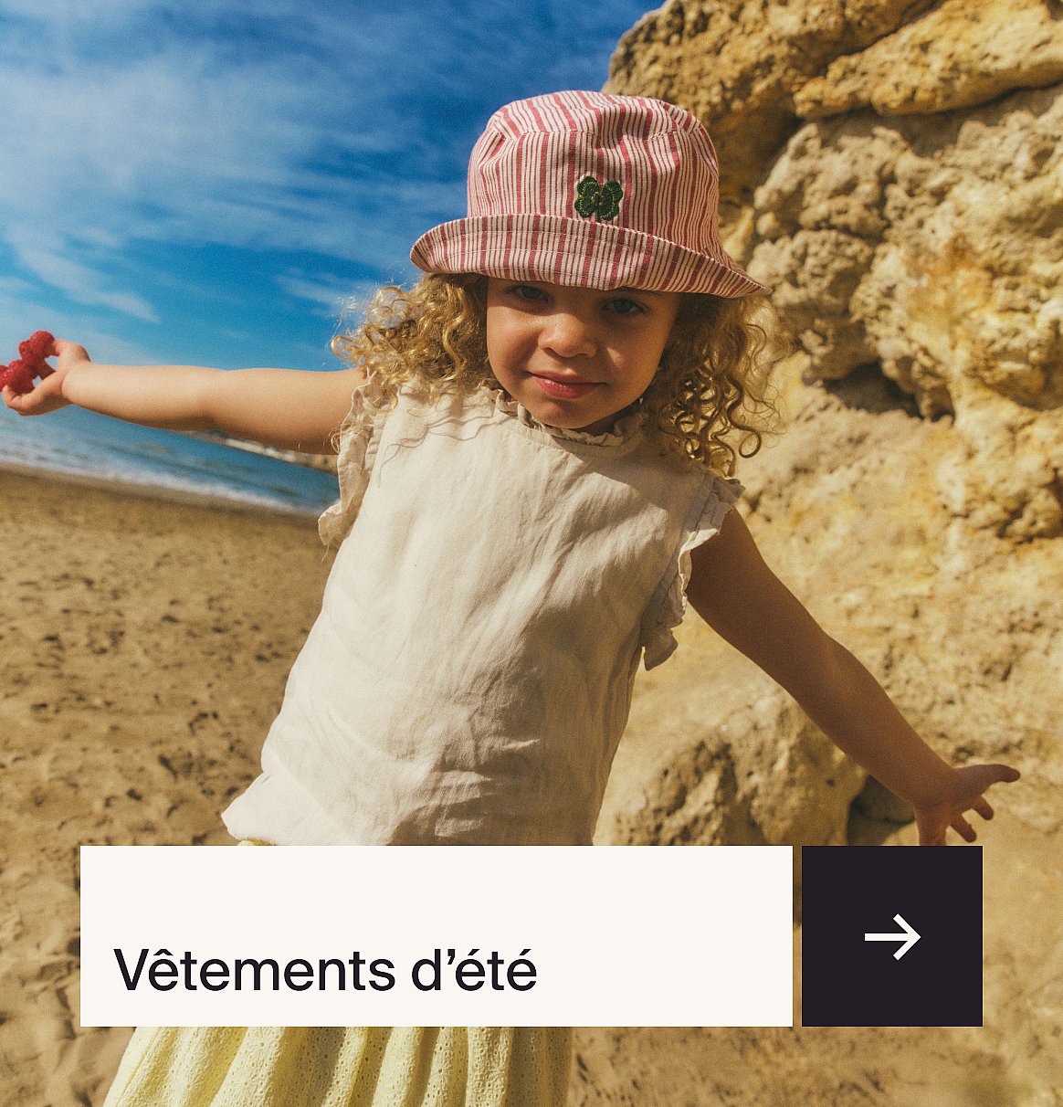 Young child in striped hat holding red berries, standing on a sandy beach by the ocean and rocky cliffs.