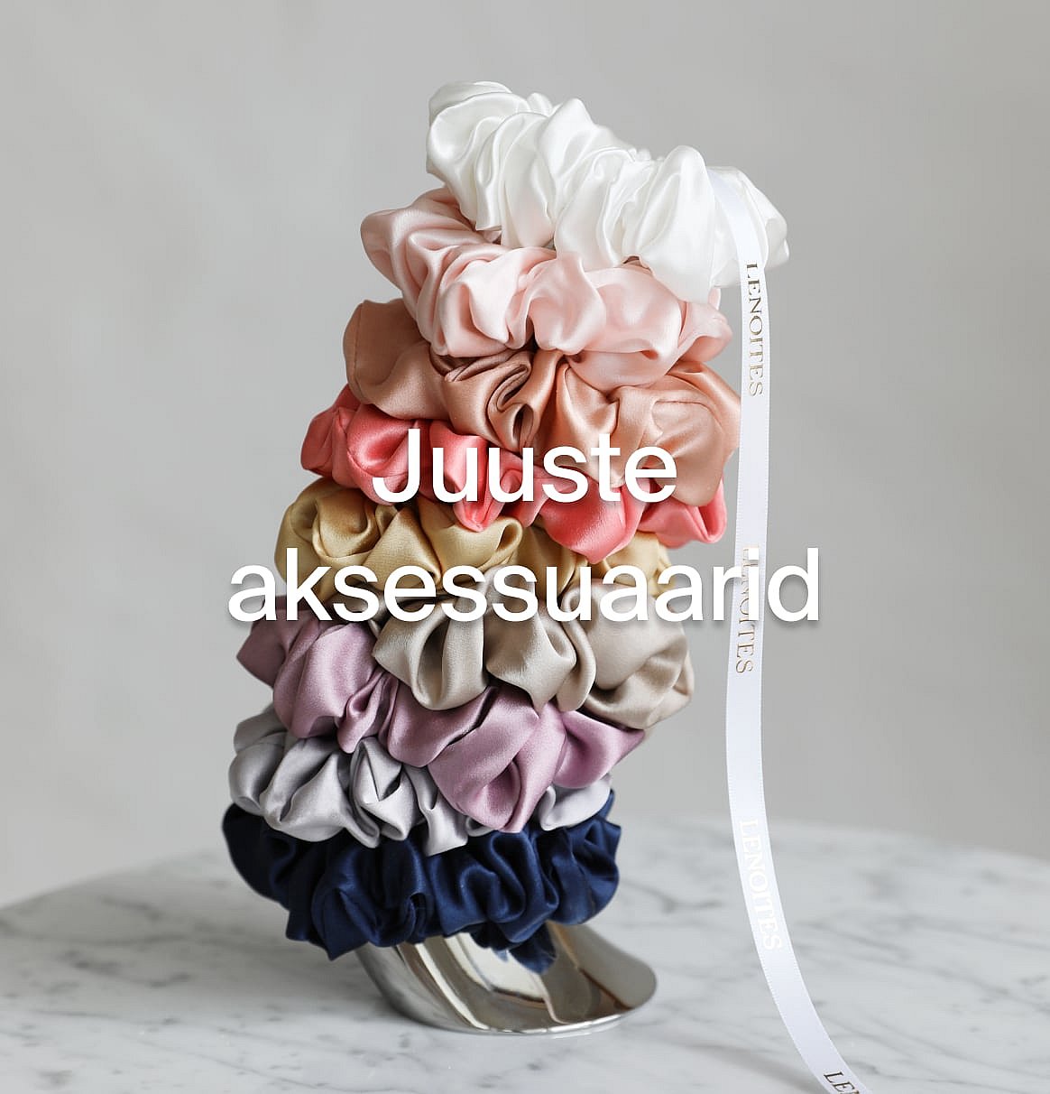 Mitmevärvilised scrunchie'd virnas marmorpinnal, valge lindiga. Tekst: Juukseaksessuaarid.