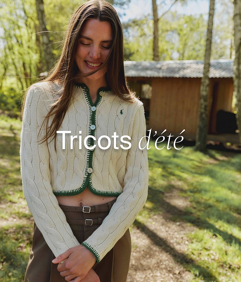 Femme souriante portant un cardigan en maille torsadée crème avec liseré vert, dans une forêt avec une cabane.
Maille d'été