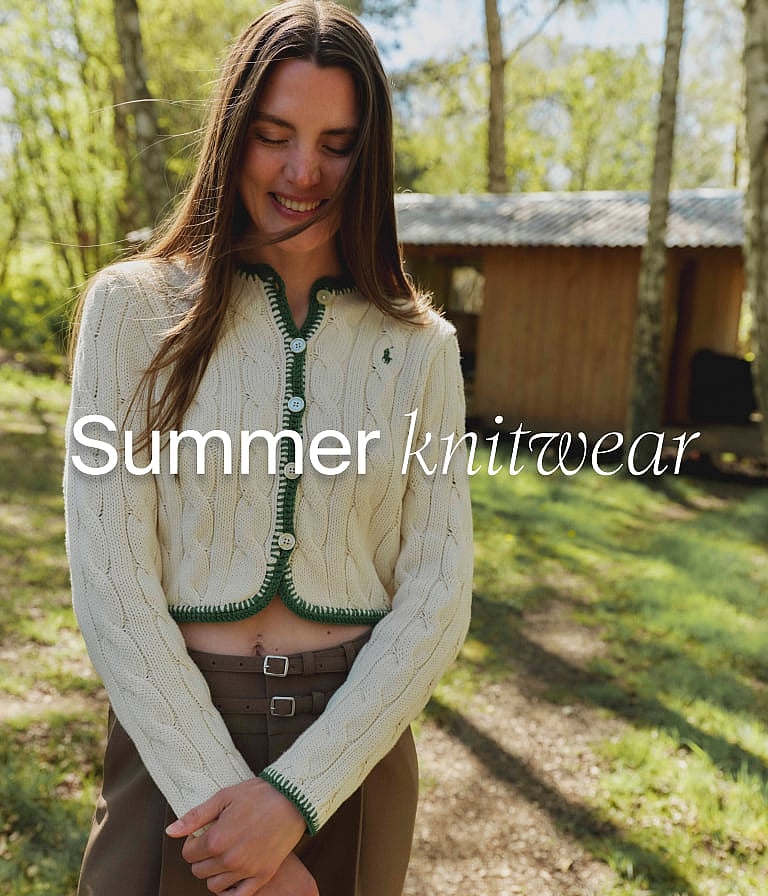 Smiling woman in a cream cable-knit cardigan with green trim, in a forest with a cabin. "Summer knitwear" text.