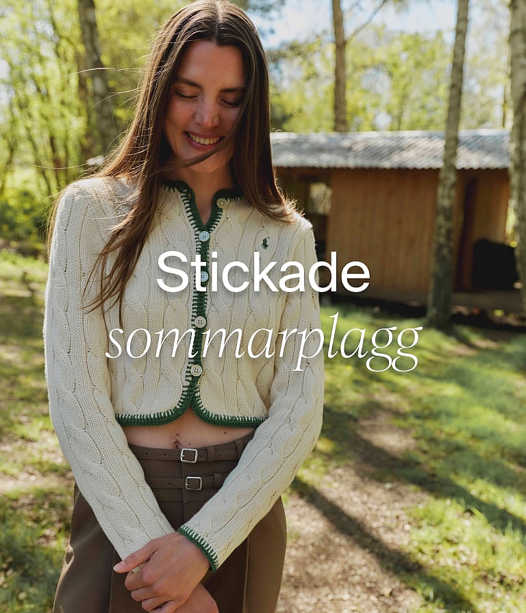 Leende kvinna i en krämfärgad flätstickad kofta med grön kant, i en skog med en stuga. "Sommarstickat" text.