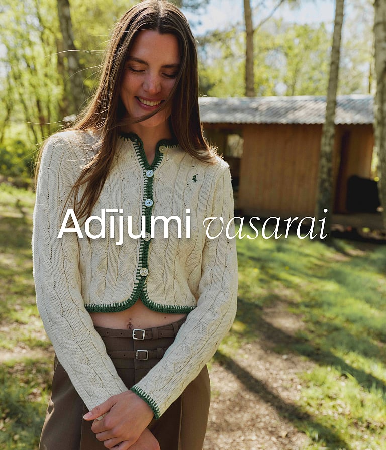 Smaidīga sieviete krēmkrāsas kabeļadījuma jakā ar zaļu apmali, mežā ar namiņu. "Vasaras adījumi" teksts.