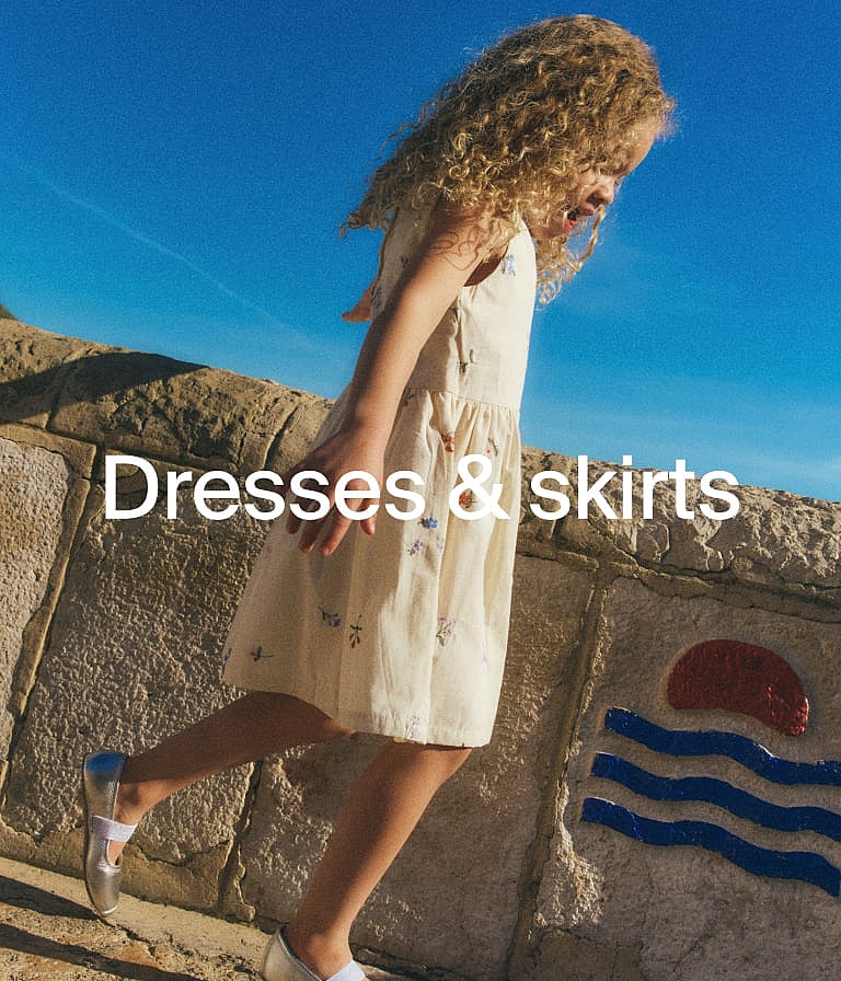 Curly-haired girl in a floral dress and silver shoes walking by a stone wall under a blue sky. Text: Dresses & skirts.
