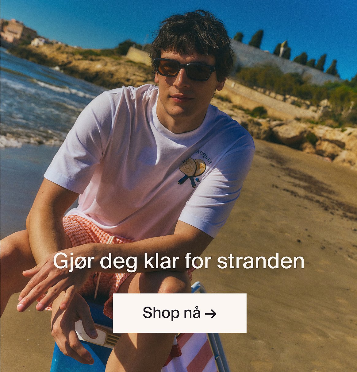 Person i hvit kortermet skjorte med hullbroderi og blå jeans, hender i lommene, står mot en lys trevegg.