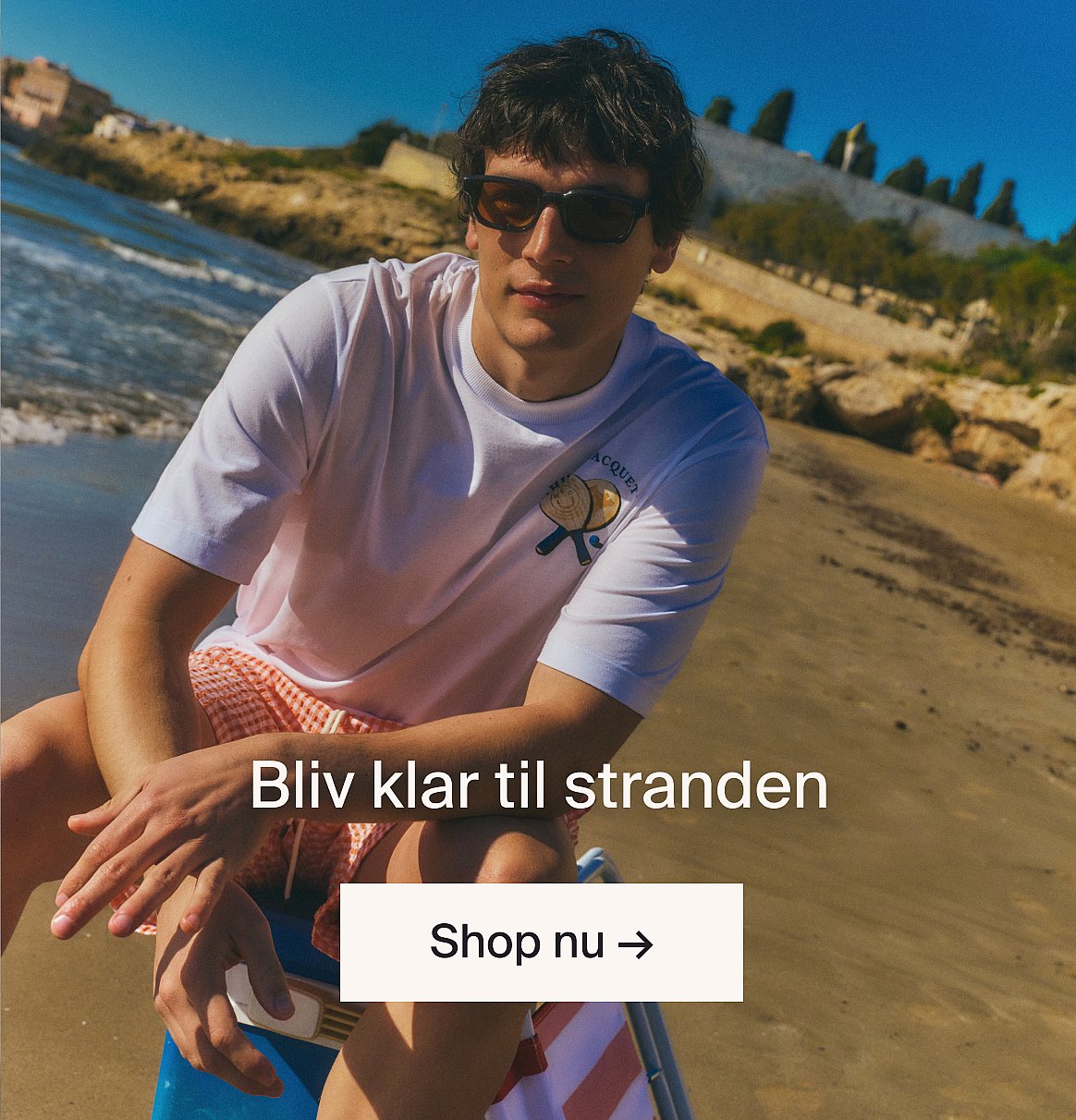 Hvid kortærmet skjorte med broderie anglaise og blå jeans, hænderne i lommerne, stående op ad en lys trævæg.