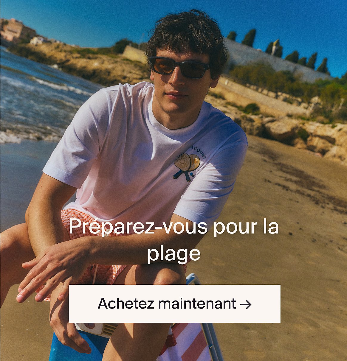 Personne en chemise blanche à manches courtes en broderie anglaise et jean bleu, les mains dans les poches, debout contre un mur en bois clair.