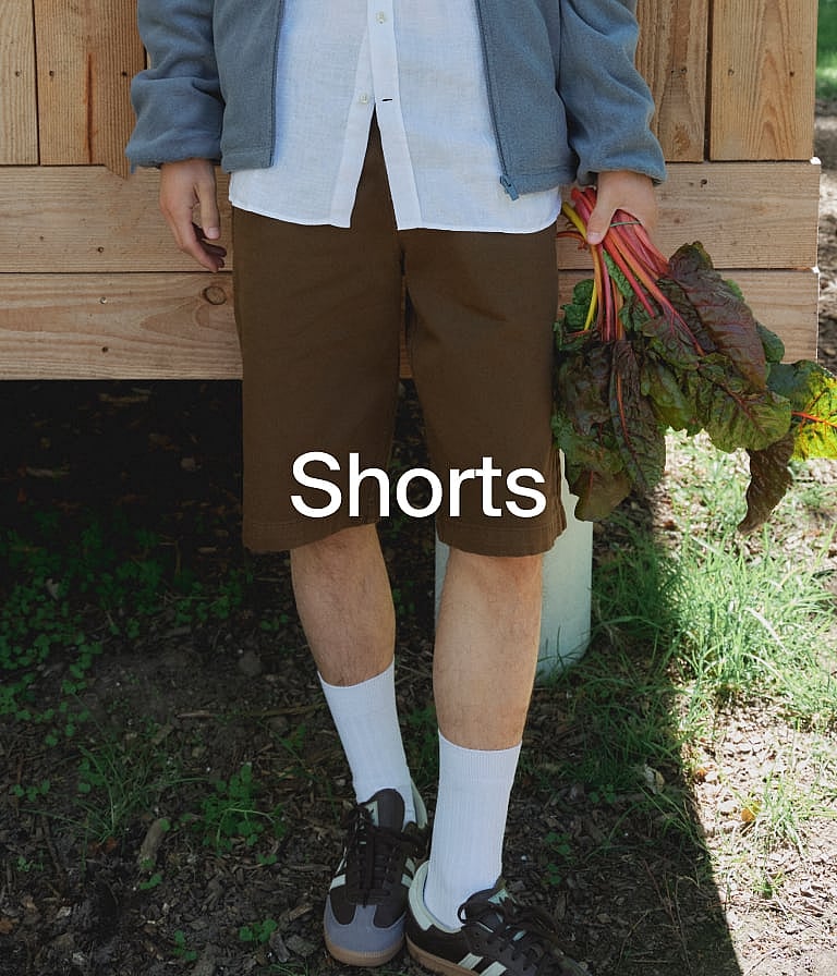 En person i brune shorts, hvid skjorte og blå jakke holder rødstilkede planter udendørs ved en trævæg.
