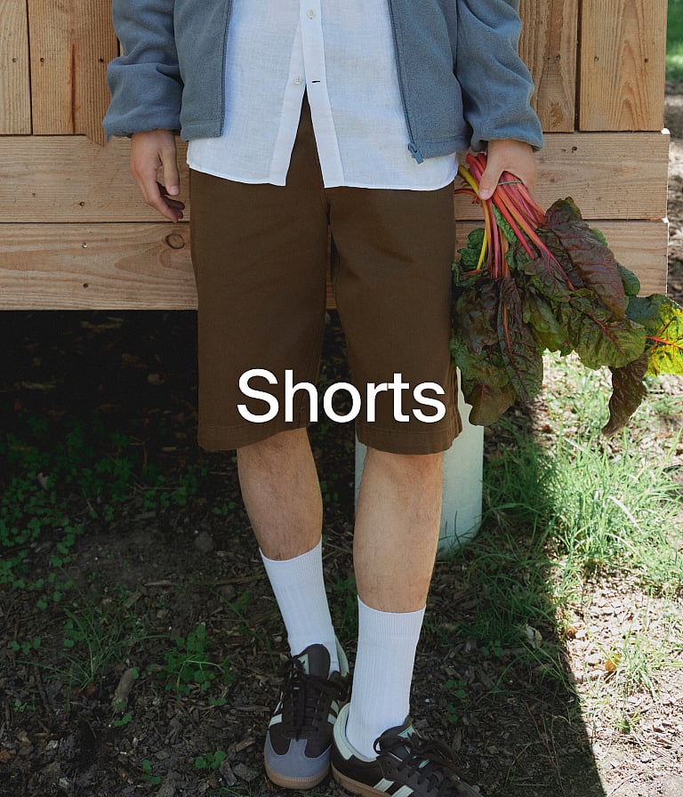 Eine Person in braunen Shorts, weißem Hemd und blauer Jacke hält rotstielige Pflanzen im Freien an einer Holzwand.