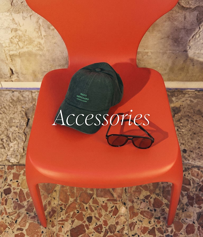 "Accessories" text over a dark green cap and black sunglasses on a bright orange chair. Stone wall and patterned floor.