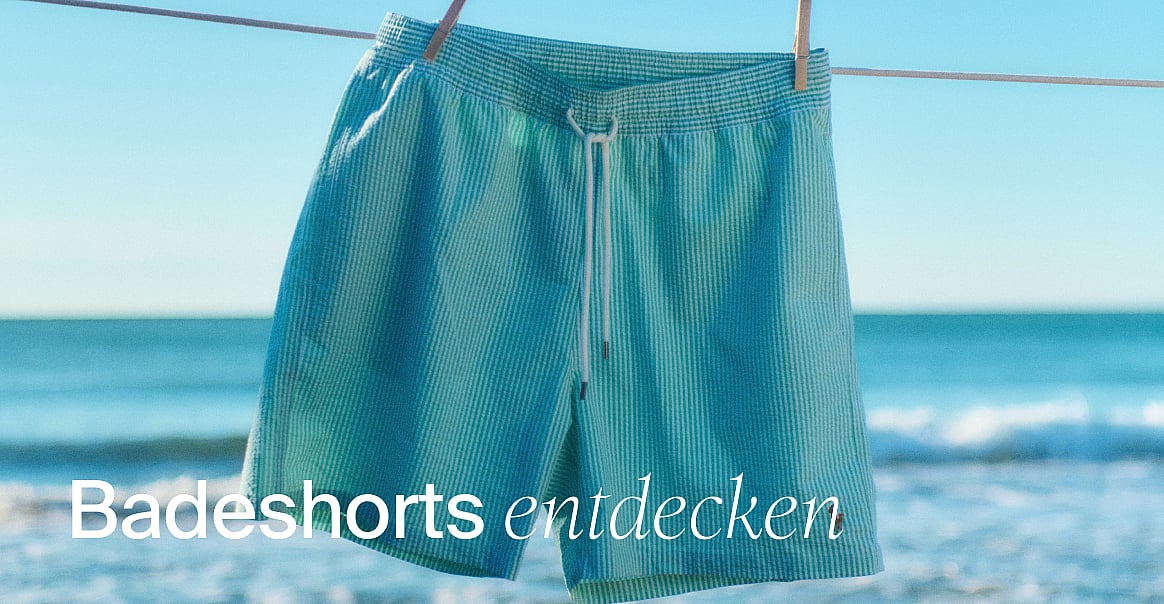 Türkis gestreifte Badeshorts hängen an einer Wäscheleine, mit einem sonnigen Strand und Meer im Hintergrund. Text: Badeshorts shoppen.