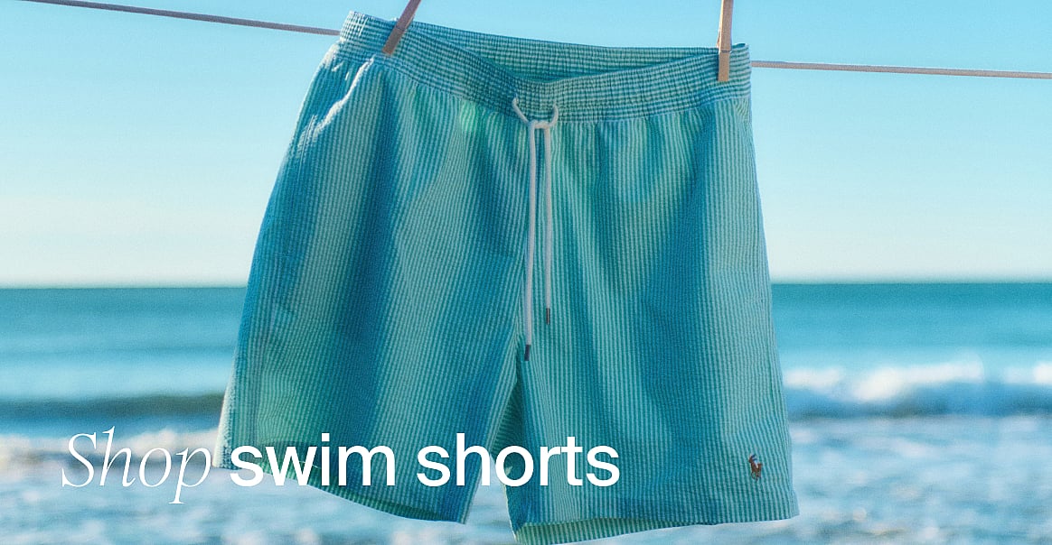 Teal striped swim shorts hang on a clothesline, with a sunny beach and ocean in the background. Text: Shop swim shorts.