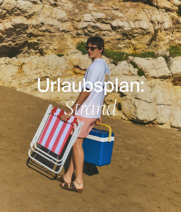 Am Sandstrand trägt eine Person einen gestreiften Strandstuhl und eine blaue Kühlbox. Im Hintergrund erheben sich felsige Klippen.
