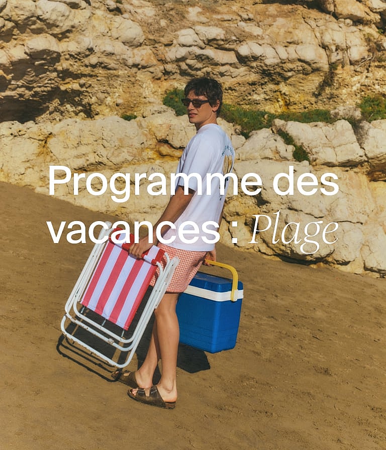 Une personne sur une plage de sable portant une chaise de plage rayée et une glacière bleue. Des falaises rocheuses en arrière-plan.