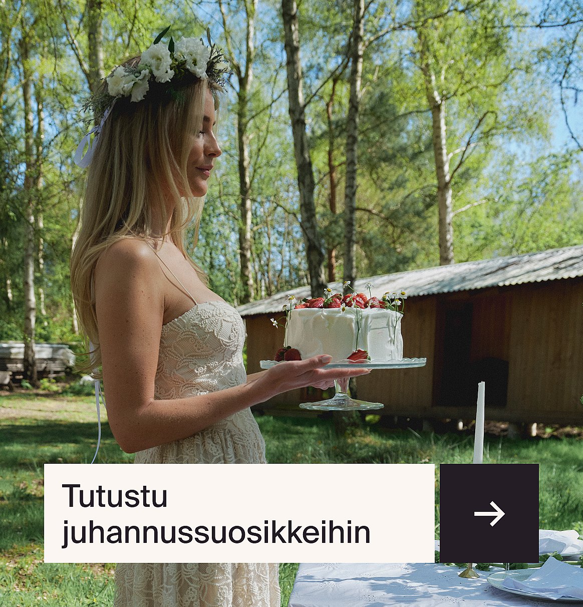 Blonde woman in flower crown and light dress holds a white strawberry cake outdoors. Sunny, green trees and a cabin are