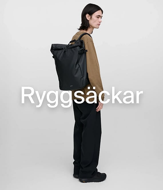En person med en svart rulltoppsryggsäck och vardagskläder, står i profil mot en enfärgad bakgrund. Ryggsäckar.