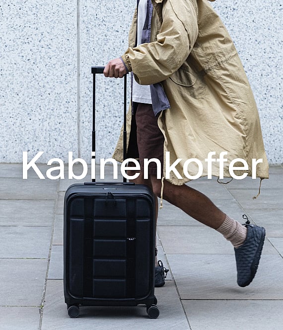 Eine Person in einem beigen Mantel zieht einen schwarzen Kabinenkoffer auf einem gepflasterten Weg. Handgepäck.