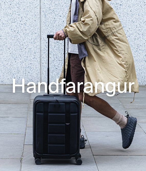 Manneskja í ljósbrúnni kápu togar svarta handfarangurstösku á malbikuðum stíg.
**Handfarangurstöskur.**