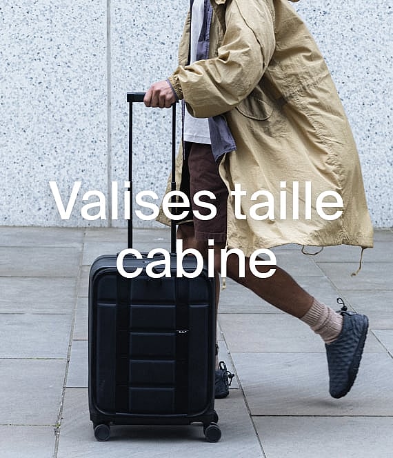 Une personne vêtue d'un manteau beige tire une valise cabine noire sur un chemin pavé.
Bagages cabine.