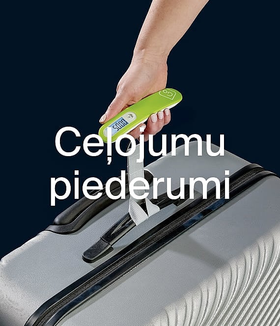 Roka tur zaļus digitālos bagāžas svarus pie pelēka čemodāna. Teksts: Ceļojumu aksesuāri.
