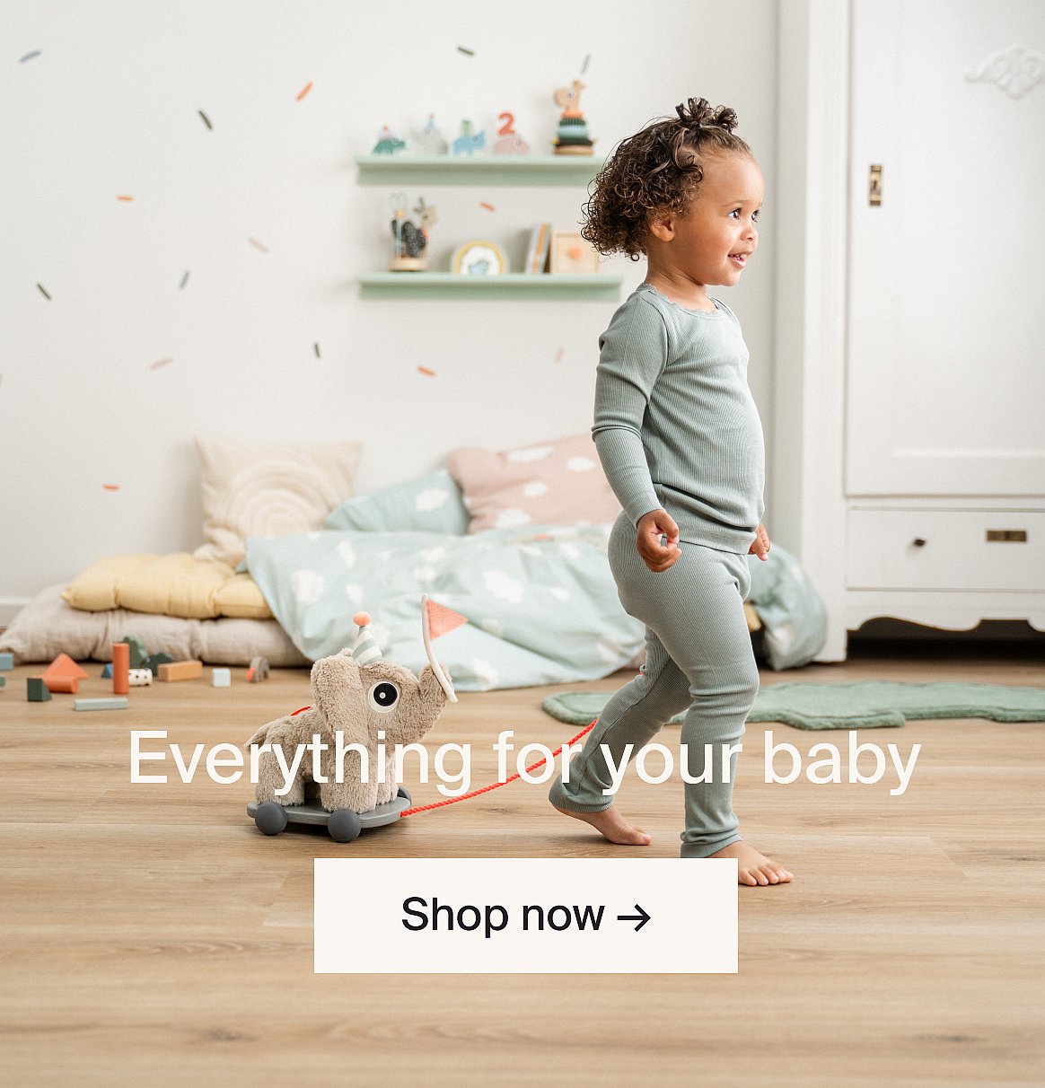 Toddler in green pajamas walking barefoot in a child's room, pulling an elephant pull-toy.