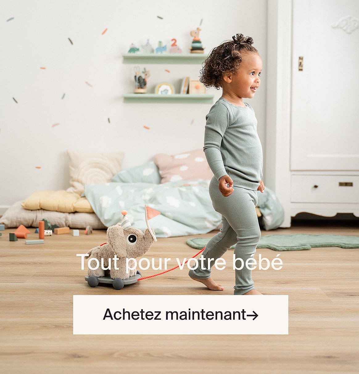 Smiling toddler with curly hair in pajamas walks across a child's room, pulling an elephant toy on wheels.
