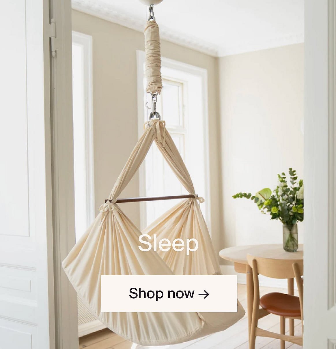 A cream fabric baby swing hangs in a bright room with a light wood table and chair.