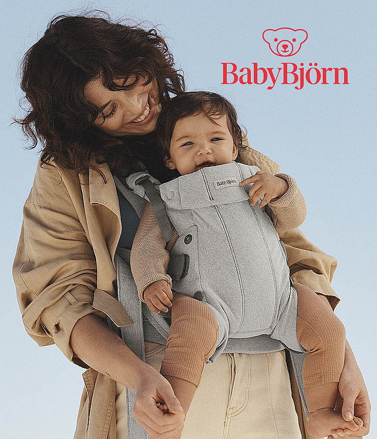 Smiling woman carries a baby in a grey BabyBjörn carrier against a clear blue sky.