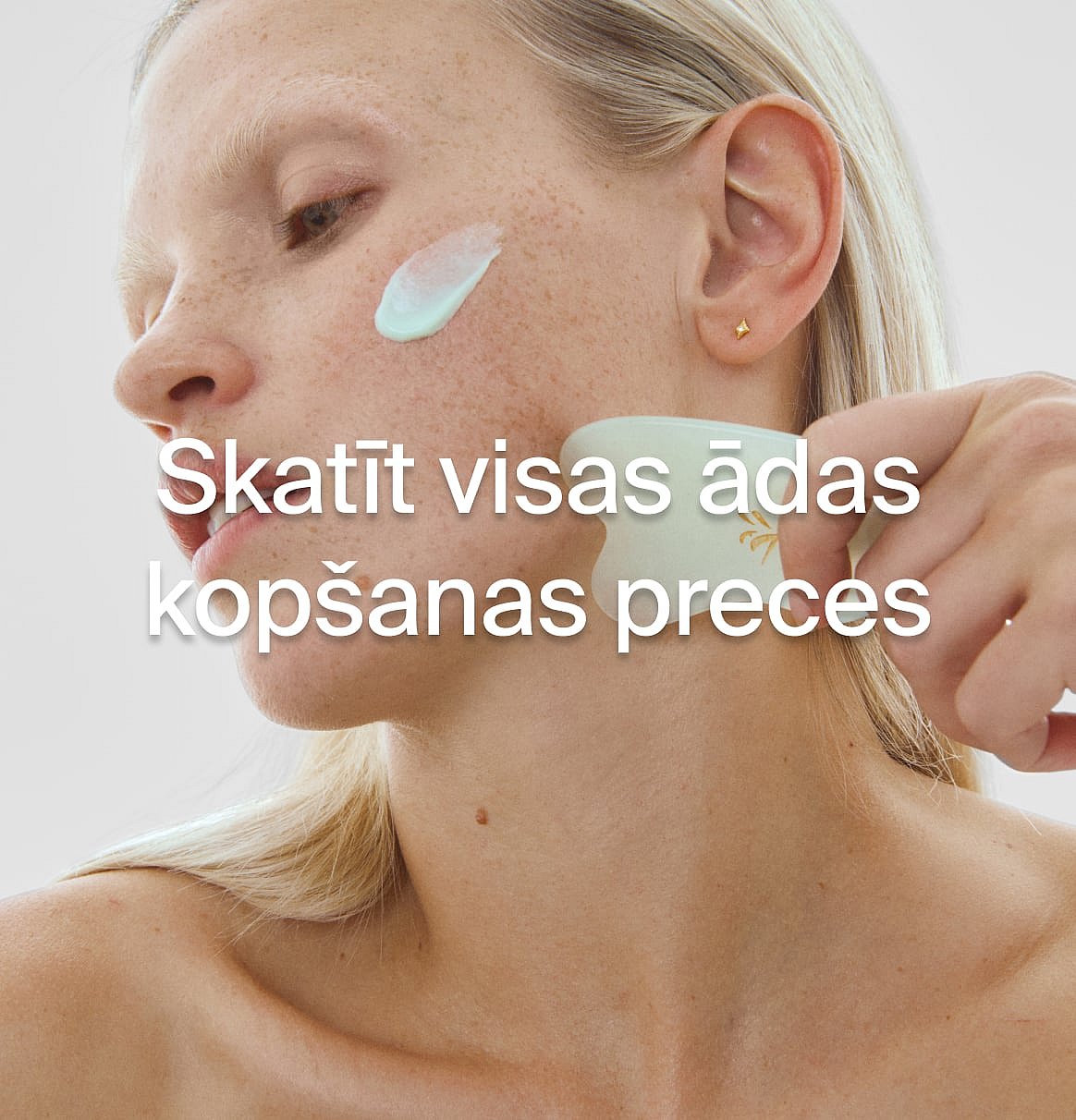 Blonda persona uzklāj krēmu uz vaiga un izmanto gua sha rīku uz kakla. Teksts: Skatīt visus ādas kopšanas produktus.