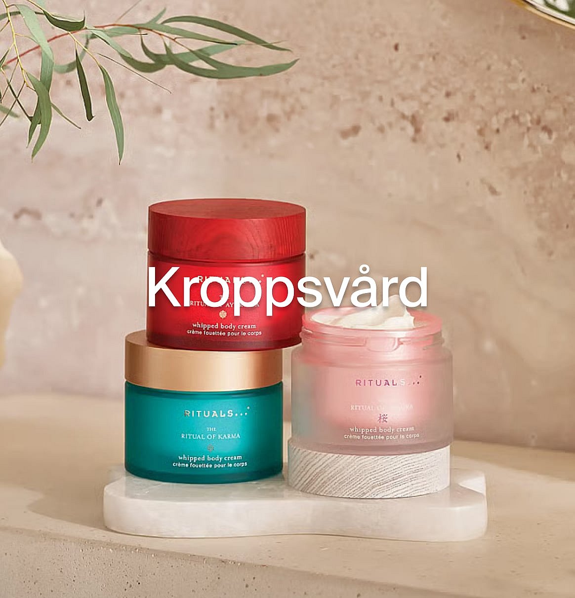 Tre Rituals kroppskrämsburkar (röda, turkosa, rosa) på en vit yta med eukalyptusblad. Text: "Kroppsvård".