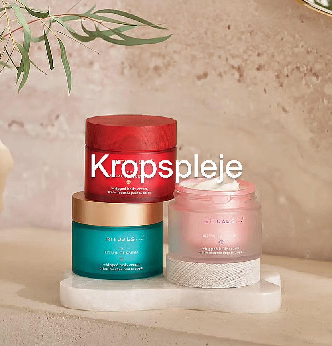 Tre Rituals kropscremekrukker (rød, turkis, pink) på en hvid overflade med eukalyptusblade. Tekst: "Kroppsvård".