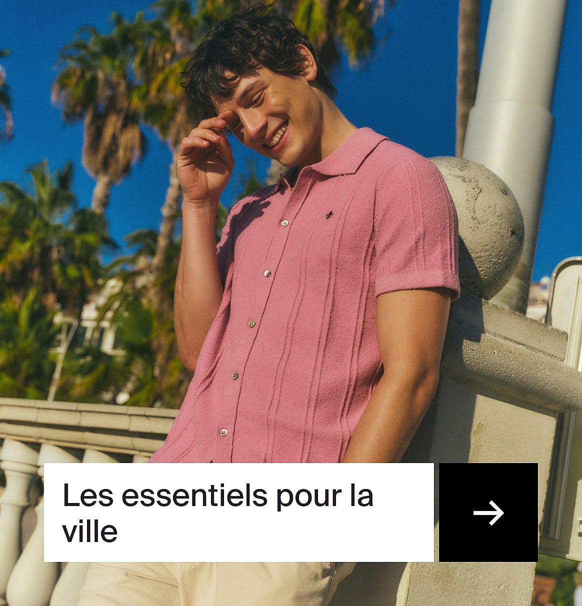 Homme souriant en polo rose, main près du visage, s'appuyant sur une balustrade en pierre. Palmiers et ciel bleu.