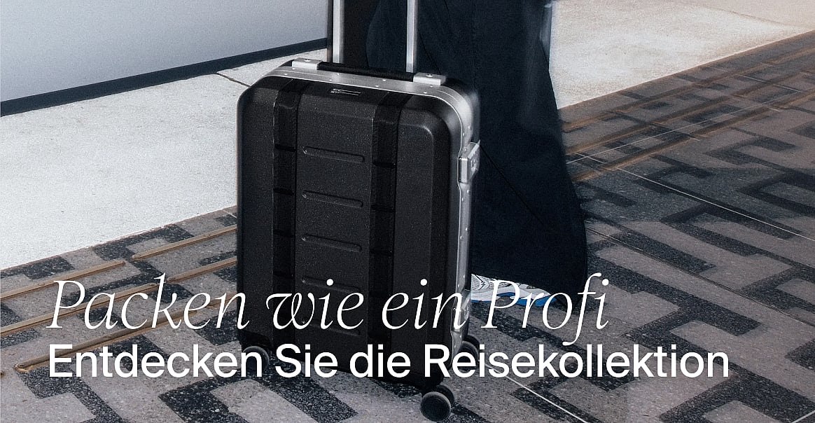 Schwarzer Rollkoffer auf einem gemusterten Boden, neben dem Bein einer Person. Text: „Packe wie ein Profi. Entdecke die Reisekollektion.“