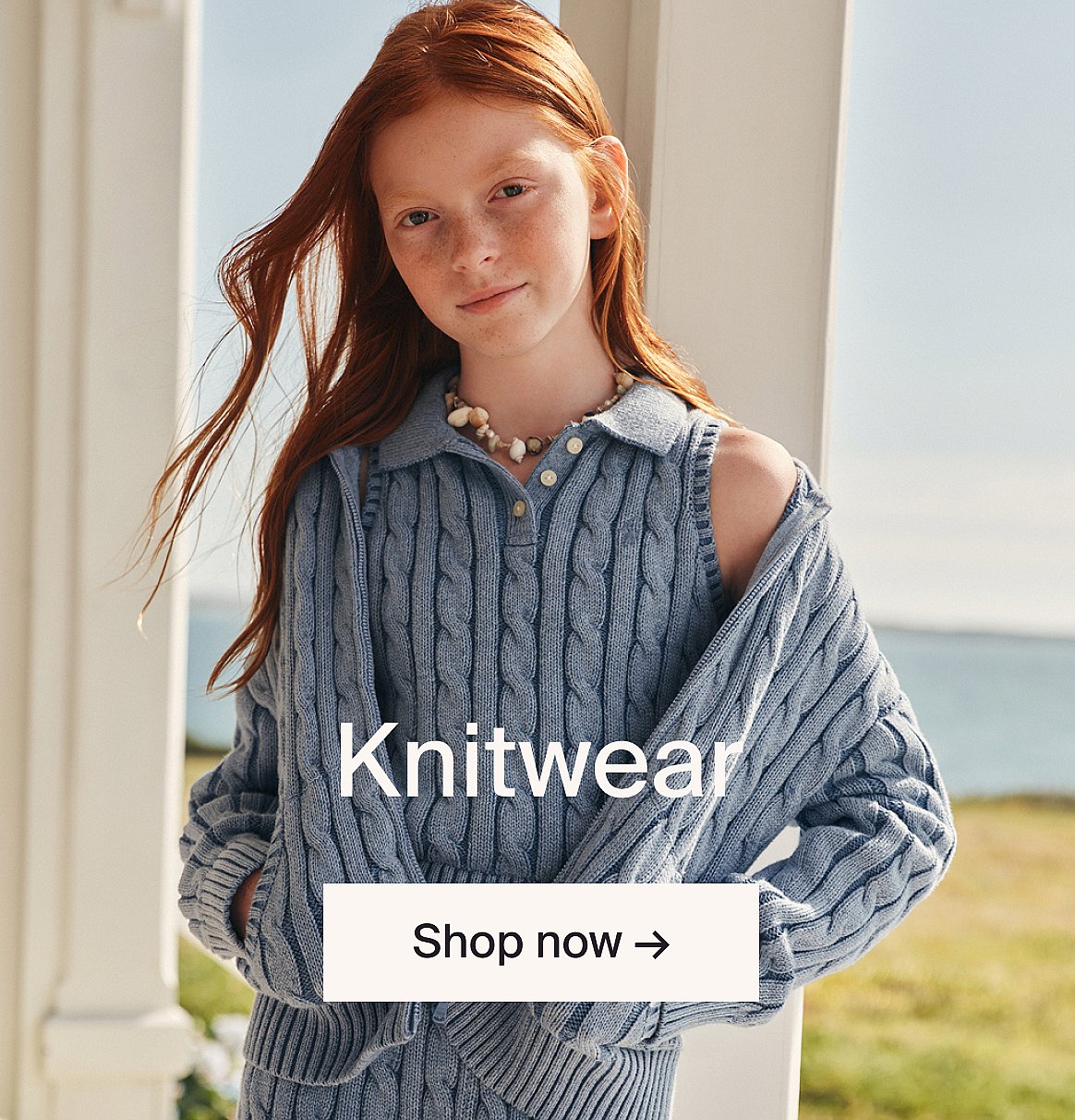 Red-haired girl in blue cable-knit sweater and vest, seashell necklace, outdoors by white columns. Text: Knitwear.